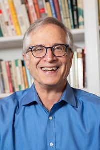 Image is of author wearing blue button up collared shirt, standing in front of bookcase